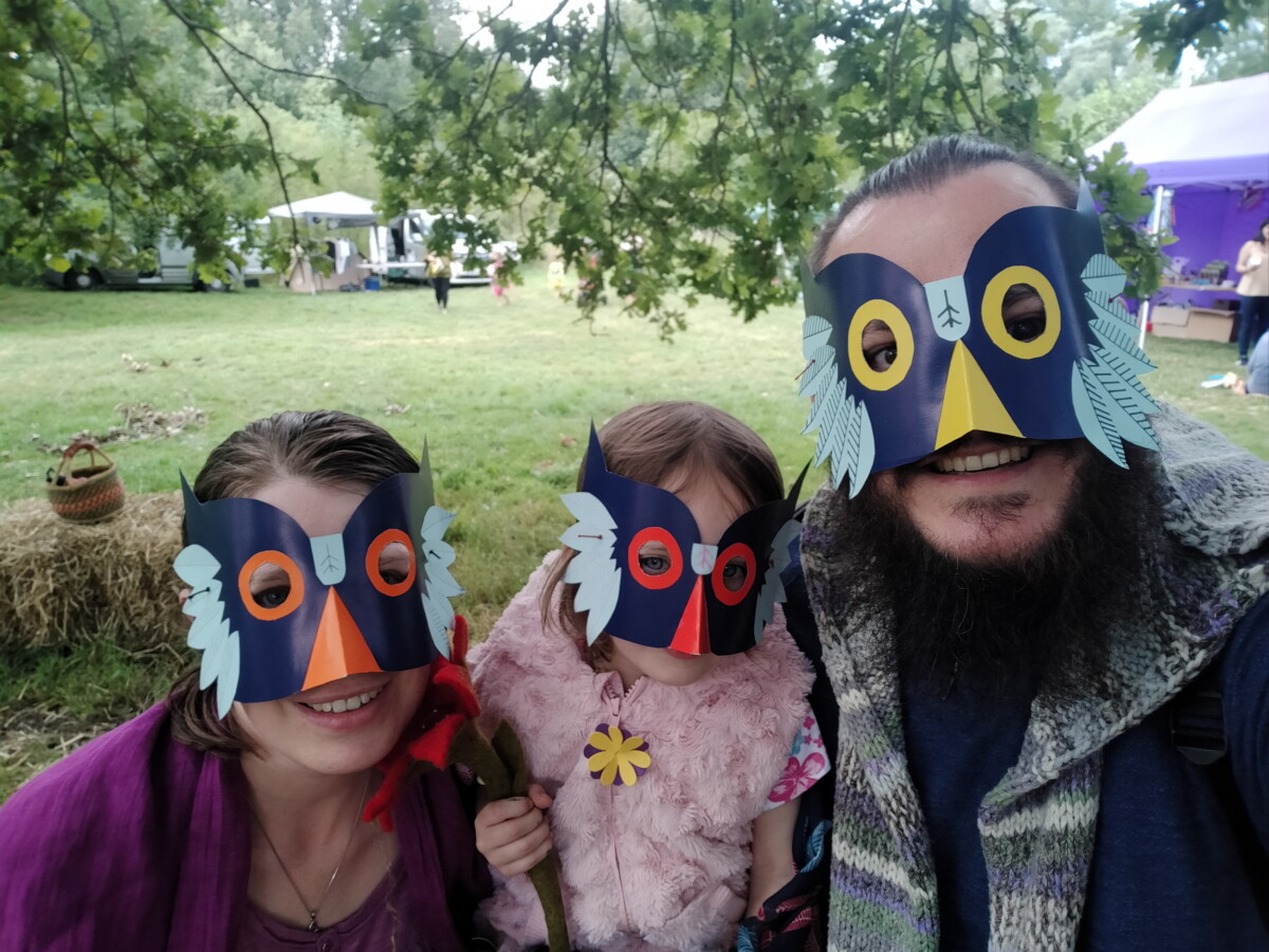 A selfie of a woman, small child and a man stood under a tree wearing home-made owl masks made from blue card with brightly-coloured additions.